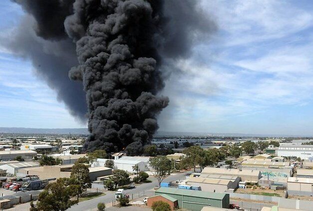 A large plume of dense, black smoke rises into the sky from an industrial complex, suggesting a significant fire and potential business interruption. The surrounding buildings appear to be warehouses or factories. The sky is mostly clear with scattered clouds. Trees and industrial units are visible.