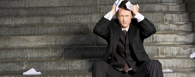 A man in a business suit sits on outdoor steps, holding his head in frustration, surrounded by crumpled papers. The grey stone surroundings add to the bleak atmosphere as he contemplates the impacts of professional indemnity on his stressful situation.