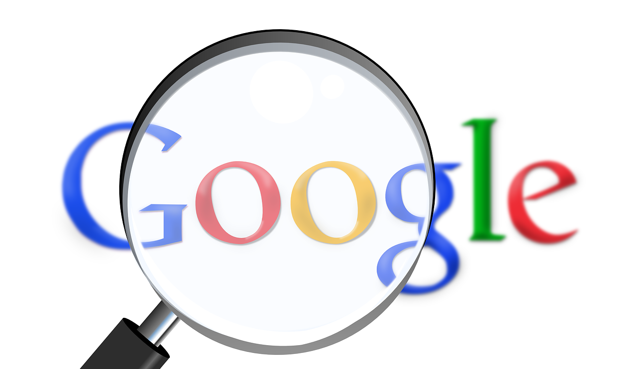 A magnifying glass is placed over the Google logo, causing the logo to appear larger and slightly blurred, with the letters in blue, red, yellow, and green. The background is white, and the magnified portion of the logo retains its original color scheme. Amidst concerns of Gmail being hacked, security awareness is heightened.