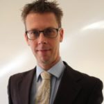A man with short, spiky brown hair and glasses is wearing a dark suit, light blue shirt, and a paisley tie. He is smiling slightly and standing against a plain, light-colored background, exuding confidence akin to one well-versed in professional indemnity insurance.