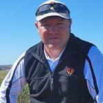 A man wearing a white cap, sunglasses on his head, and a black and white jacket stands in a field with crops. He is looking at the camera with a slight smile, perhaps contemplating the peace of the clear blue sky behind him. His confident stance suggests he understands the importance of professional indemnity insurance for his work.