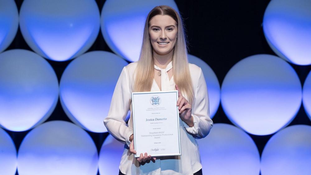 A person with long hair wearing a light blouse poses with a certificate against a backdrop of large, illuminated circular panels. Jessica Dametto looks content and stands centered in the frame, showcasing the certificate prominently.