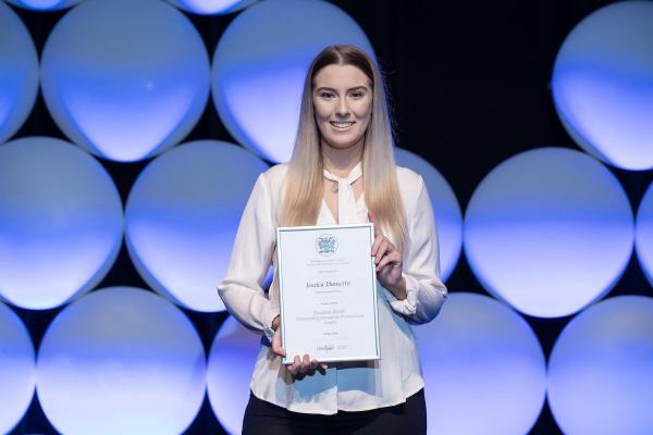 A person with long hair wearing a light blouse poses with a certificate against a backdrop of large, illuminated circular panels. Jessica Dametto looks content and stands centered in the frame, showcasing the certificate prominently.