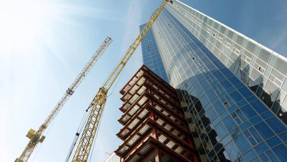 A high-rise building under construction, overseen by an accredited professional with NSW Building Designer Registration, stands tall against a clear, sunny sky. Yellow cranes tower above, and the building's glass facade and visible steel framework gleam as sunlight streams in from the top left corner.