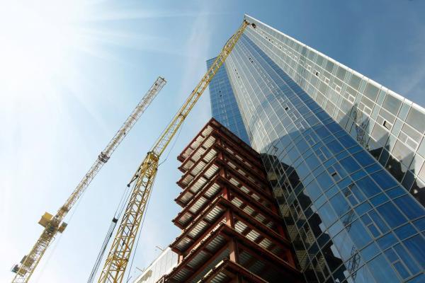 A high-rise building under construction, overseen by an accredited professional with NSW Building Designer Registration, stands tall against a clear, sunny sky. Yellow cranes tower above, and the building's glass facade and visible steel framework gleam as sunlight streams in from the top left corner.