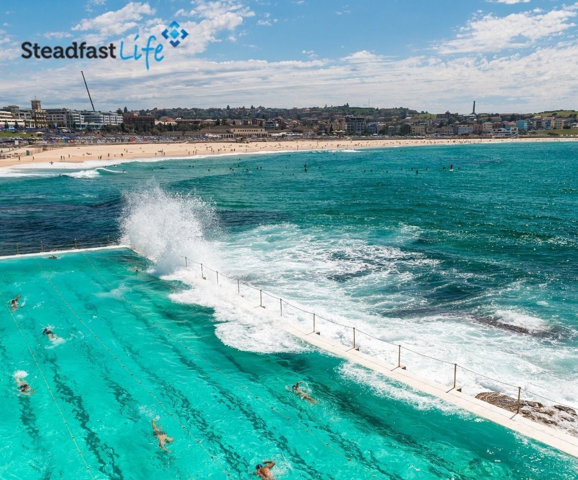 Steadfast Life A beachfront scene welcomes you with a vibrant turquoise ocean and an outdoor swimming pool adjacent to the sea. Waves crash over the pool's edge as people swim. In the background, there is a sandy beach with visitors and a distant urban skyline. The text "Steadfast Life" is visible.