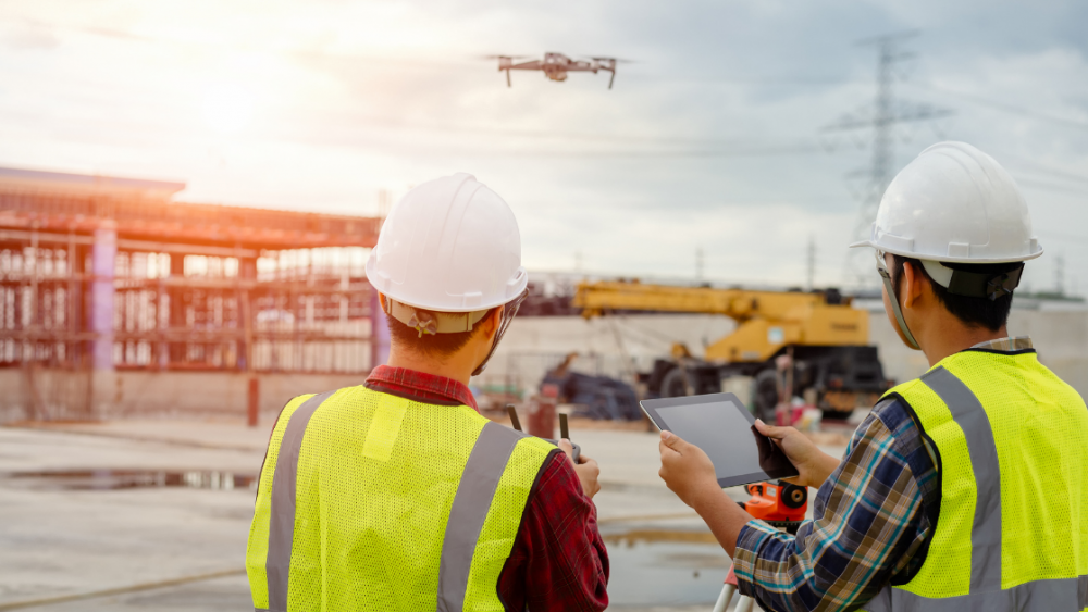 Two construction workers in high-visibility vests and helmets use a tablet and a remote control to operate a flying drone at a construction site. The background shows partially constructed buildings and construction equipment under a cloudy sky, highlighting business use of drones for site management.