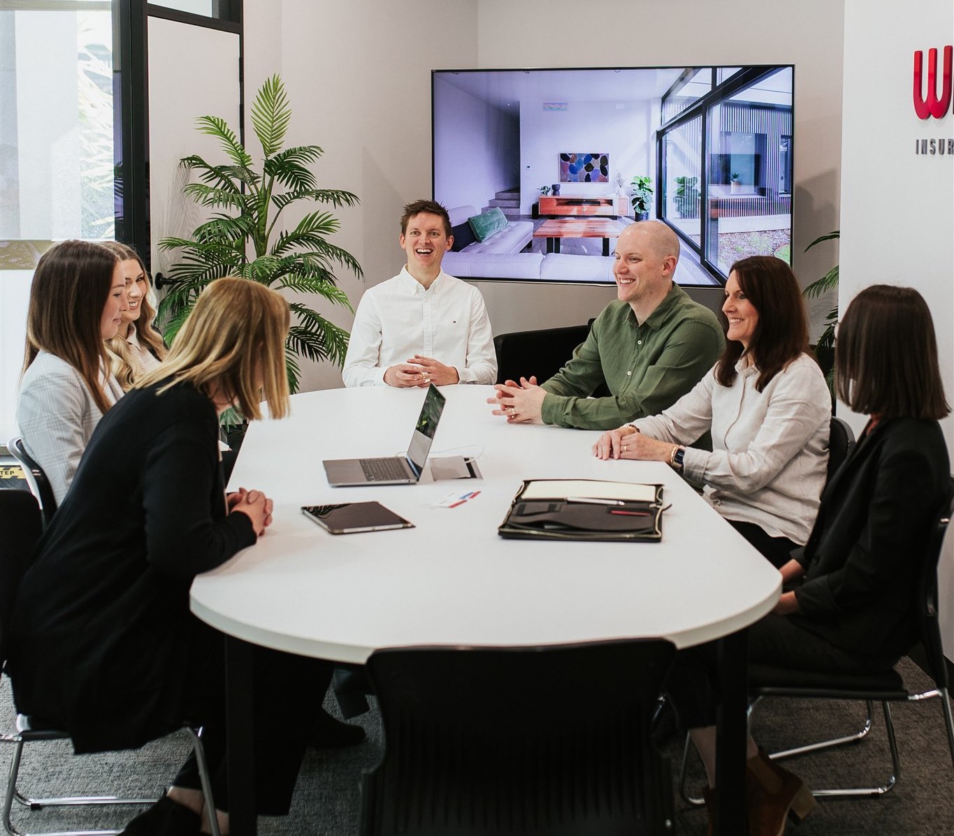 A group of six colleagues, four women and two men, are seated around a white table for a meeting in a modern office. They are engaged in conversation and appear to be smiling. A large screen displaying a room interior is visible in the background, with potted plants in the corners, fostering an environment ripe with job opportunities.